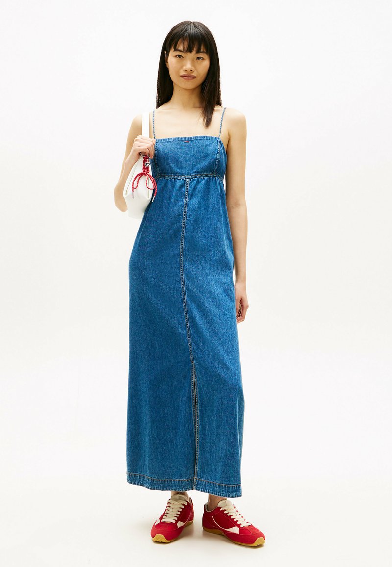 Kobieta w długiej niebieskiej sukience dżinsowej, czerwono-białych trampkach, trzymająca białą torebkę z czerwonymi detalami na jednolitym białym tle.