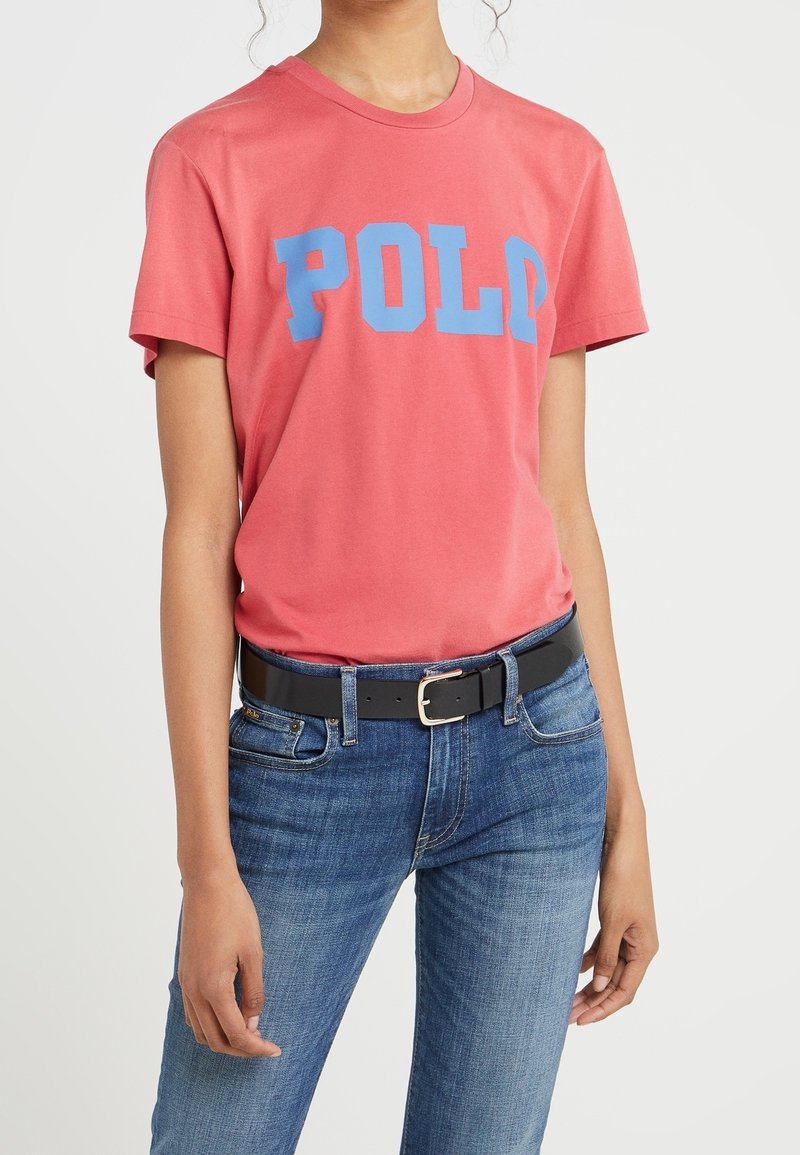 Červené bavlněné tričko s modrým nápisem "POL", spárované s modrými džínovými kalhotami a černým opaskem se stříbrnou sponou. Krátké rukávy a kulatý výstřih.