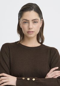 Pruun kootud sviiter ümarate kaelustega, millel on tekstureeritud kangas ja kuldsed nupud varruka äärisel. Modelli käed on rüpes.