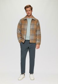 Veste en plaid marron et gris avec un t-shirt bleu, associée à un pantalon gris et des baskets blanches ; la veste présente un col et une texture douce.
