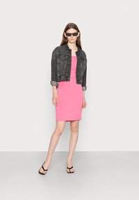 Vestido justo rosa combinado com uma jaqueta de ganga preta, com mangas enroladas. Modelo usa óculos de sol retangulares e chinelos pretos.