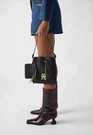 Sac seau noir en tissu tissé avec un cordon de serrage, un logo doré en accent et une bandoulière en cuir. Assorti à des bottes à talons hautes jusqu'au genou de couleur marron foncé.
