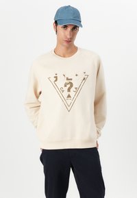 Felpa beige con un logo triangolare marrone e crema, con maniche raglan e scollatura rotonda. Abbinata a pantaloni blu navy e un cappellino di denim.