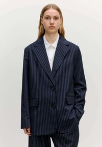 Marineblaues gestreiftes Blazer, strukturierte Form mit geknoteten Revers, zwei Fronttaschen und Zwei-Knopf-Verschluss. Glatte Stofftextur.