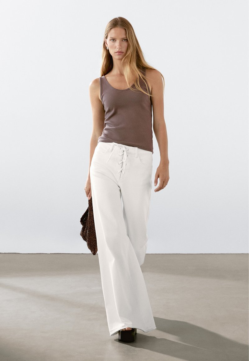Witte wijde broeken met een vetersluiting aan de voorkant, in combinatie met een strakke bruine tanktop. Het model draagt een gestructureerde donkere tas en heeft chunky hakken aan.