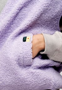 Χέρι μέσα σε τσέπη γκρι μανικιού, που φοράει απαλό ροδακινί ένδυμα από μαλακό, σγουρό φλις με μικρή μαύρη και κίτρινη ετικέτα "PCS".