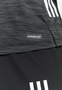 Tummanharmaa tekstuurilla varustettu urheiluvaatemateriaali, jossa on valkoinen "AEROREADY" -teksti ja kolme valkoista raitaa yläreunan lähellä.