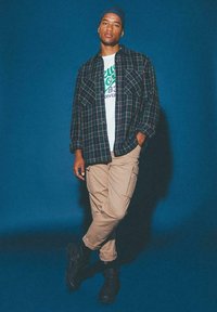 Koszula flanelowa w czarno-zieloną kratę, biała koszulka z zielonym napisem, khaki spodnie cargo i czarne buty wojskowe. Niebieskie tło.