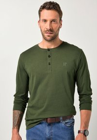 Mies, joka käyttää oliivinvihreää pitkähihaista henley-paitaa, jossa on kolme nappia, sinisiä farkkuja, ruskeaa vyötä, ruskeaa kelloa ja tatuointia vasemmassa kyynärvarressa.