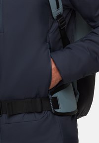 Persona con giacca blu scuro, mano nella tasca laterale e tracolla blu chiaro con fibbia nera in vita.