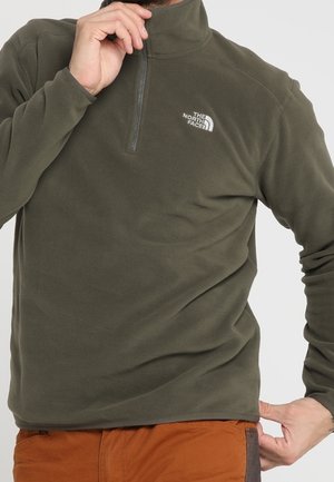 Mężczyzna w oliwkowo-zielonej polaru North Face z przednim zamkiem, regulujący kołnierz, w połączeniu z pomarańczowymi spodniami.