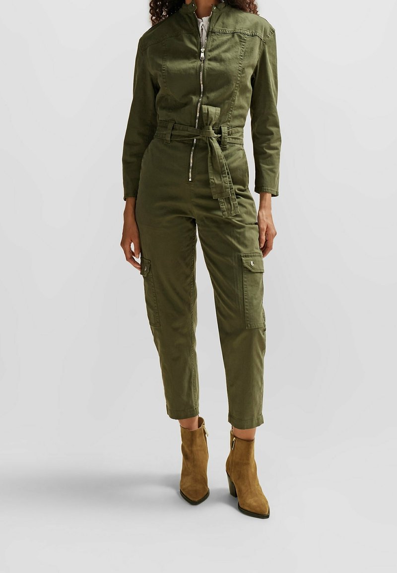 Femme portant une combinaison utilitaire vert olive avec fermeture éclair devant, lien à la taille, poches cargo et bottines en daim marron sur fond uni.
