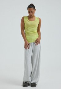 Mujer con cabello oscuro recogido en un moño, vistiendo una camiseta sin mangas amarilla y pantalones sueltos grises, de pie con las manos delante contra un fondo liso.