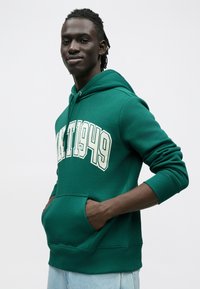 Sudadera verde con capucha y bolsillo tipo canguro, con el texto "MT1949" en color crema en el pecho. Puños y dobladillo acanalados.