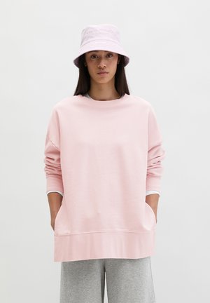 Donna che indossa una felpa oversize rosa chiaro, un cappello a secchiello abbinato e pantaloni grigi, in piedi con le mani nelle tasche davanti a uno sfondo semplice.