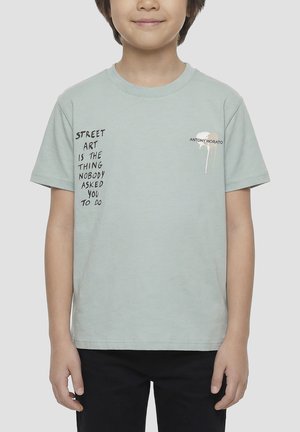 Child wearing a light green T-shirt with black text reading "Street art is the thing nobody asked you to do" and a small white paint splash logo.