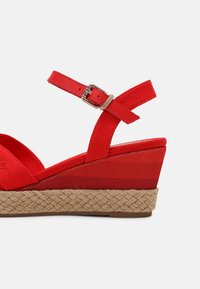 Sandale rouge à talon en forme de coin avec une sangle de cheville, boucle en argent, plateforme texturée et semelle en jute tressé sur fond blanc.