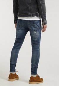 Jeans de denim azul ajustados con un lavado desgastado, bolsillos traseros y detalles de costura intrincados. Combinados con zapatos de ante marrón.