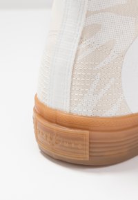Beige texturerad sneaker med meshfinish, vita accenter och gummisula. Har en rektangulär logotypbricka i orange på hälen.