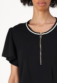 Musta lyhythihainen paita, jossa on pyöreä kaula-aukko. Paidassa on kontrastina valkoinen reunus ja kultainen vetoketju edessä. Sileä kangas.