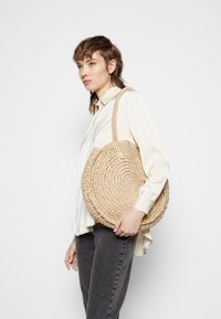 Rund vävd stråväska med två långa handtag, i en naturlig beige färg och en texturerad yta. Väskan hålls vid sidan.