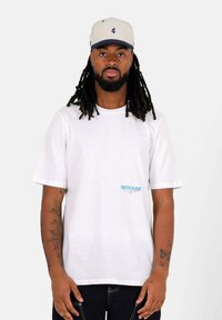 Wit katoenen T-shirt met korte mouwen, met het "SIXTH JUNE"-logo in blauw en roze. Model draagt een gestreepte pet met klep.