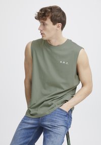 Oliven grøn ærmeløs t-shirt med rund hals og hvid trykt "A.R.C." logo, parret med blå denim jeans. Enkel design.