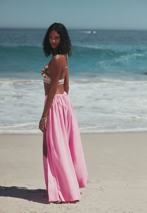 Ung kvinde står på en sandstrand og vender mod havet iført en crop top med palmeprint og en flydende lyserød nederdel med slids.