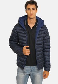 Veste matelassée bleu marine avec capuche amovible, motif matelassé horizontal, fermeture à glissière frontale et poignets élastiqués, portée sur un polo noir.