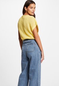 Maglietta a maniche corte di colore giallo, lavorata a maglia con trama larga e aperta. Abbinata a jeans in denim azzurro chiaro a vita alta con tasche posteriori.