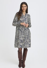 Kleid mit einem creme- und marineblauen abstrakten Muster, mit Kragen, langen Ärmeln und lockerer Passform, kombiniert mit braunen Wildleder-Stiefeln bis zum Knie.