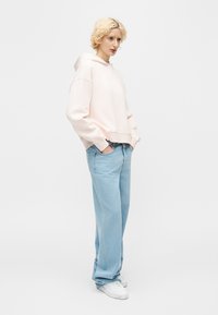 Personne aux cheveux blonds courts et bouclés, portant un sweat à capuche rose pâle, un jean large bleu clair et des baskets blanches, debout les mains dans les poches.