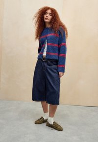 Jonge vrouw met krullend rood haar, gekleed in een blauw en rood gestreepte trui, witte top, wijde denim shorts, witte sokken en bruine schoenen.