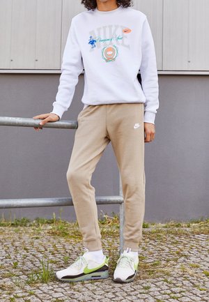Persoon die beige Nike joggingbroek draagt, wit Nike sweatshirts met meerkleurige print en witte sneakers met groene Nike swooshes, staand bij een metalen railing.