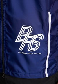 Veste bleue avec fermeture éclair blanche et bande verticale, affichant le logo "BRS" et le texte "Blue Ribbon Sports Track Club" sur le devant.