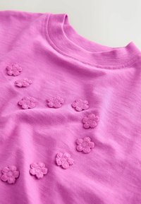 Rózsaszín pamut póló, amely texturált, hímzett virágdíszekkel van ellátva az elején. Kerek nyakkivágás és sima anyagfelület.