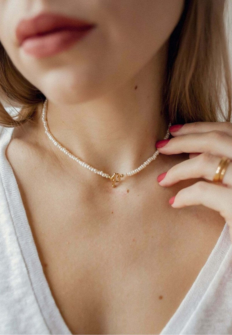 Femme portant un collier de perles avec fermoir en or, touchant le collier, montrant le cou nu et un haut blanc à col en V, vernis à ongles rose à la main.