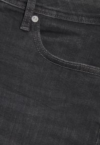 Detailní záběr na kapsu černých džínů s viditelným šitím a kovovou nýtkou v horním levém rohu.