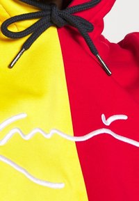 Primo piano di una felpa gialla e rossa con lacci neri e un design bianco ricamato a forma di onda che attraversa il centro.