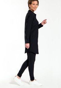 Zwarte longsleeve shirtjurk, met knopen aan de voorkant, gebogen zoom, gecombineerd met zwarte leggings en witte sneakers, wat een gestroomlijnde look benadrukt.