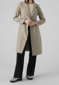 Manteau en mélange de laine beige avec un design ajusté, revers crantés, manches longues et une coupe mi-longue, associé à un pantalon noir et des chaussures argentées.