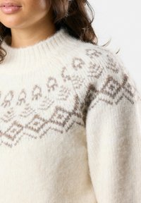 Crèmewitte sweater van getextureerde wol, met een geometrisch bruin patroon rond de hals en schouders. Chunky breiontwerp.