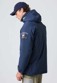 Mørkeblå vandtæt jakke med hætte, der har et broderet logo-patch på skuldren og kontrasterende manchetter. Tekstureret stof og tætsiddende design.