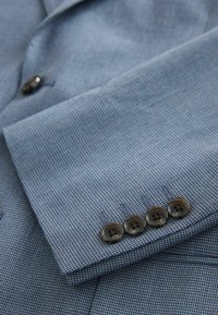 Manche de blazer bleu clair texturé avec quatre boutons foncés alignés près du poignet sur un fond en tissu assorti.