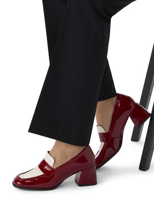Talons en cuir verni rouge avec une forme bloc, présentant un accent blanc contrastant sur le devant et un bout arrondi. Bas de pantalon noir visible.