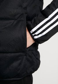 Svart vadderad jacka med tonal präglad "adidas"-logotyp. Har en dragkedjeficka och vit tre-randsdesign längs ärmen.