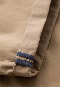 Nærbillede af beige stof med en foldet kant, fremhævet af to parallelle, mørkeblå syede bånd.