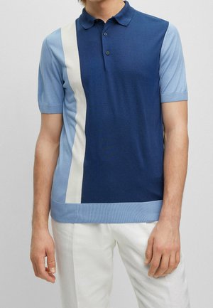 Mann trägt ein kurzärmeliges Poloshirt mit hellblauen Ärmeln, einer dunkelblauen und weißen vertikalen Streifenfront und weißen Hosen.
