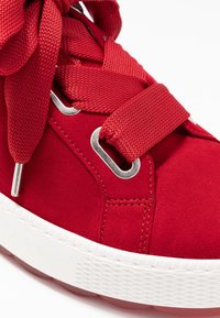 Zapatilla de gamuza roja con parte superior texturizada, grandes cordones planos, ojales de metal y una suela de goma blanca con fondo translúcido rojo.