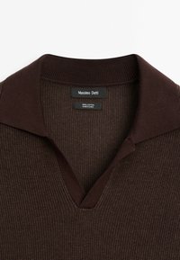 Polo en coton marron avec un design texturé, doté d'un col en V et d'un col lisse contrastant. Les étiquettes indiquent la marque et le matériau.